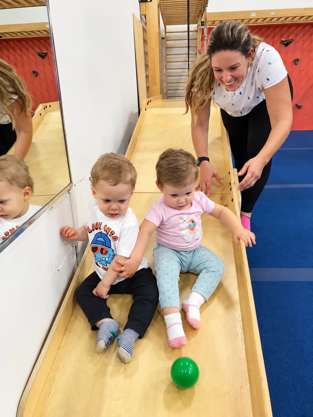 A parent laughs with two toddlers as they play on a wooden ramp in the blue gym