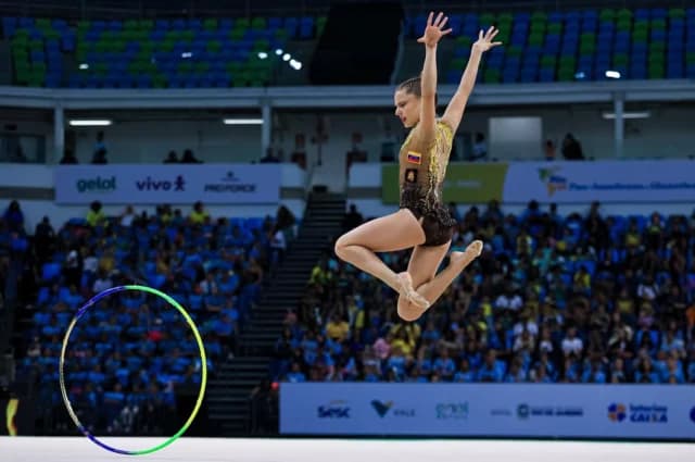 A gymnast alone on the competition carpet, mid-leap during her hoop routine, with the arena crowd behind her