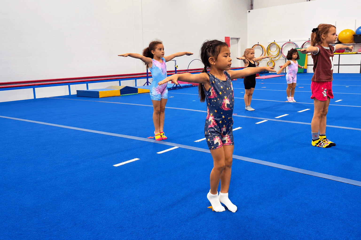 Five girls in a Level 1 gymnastics class stand with arms out on a blue floor