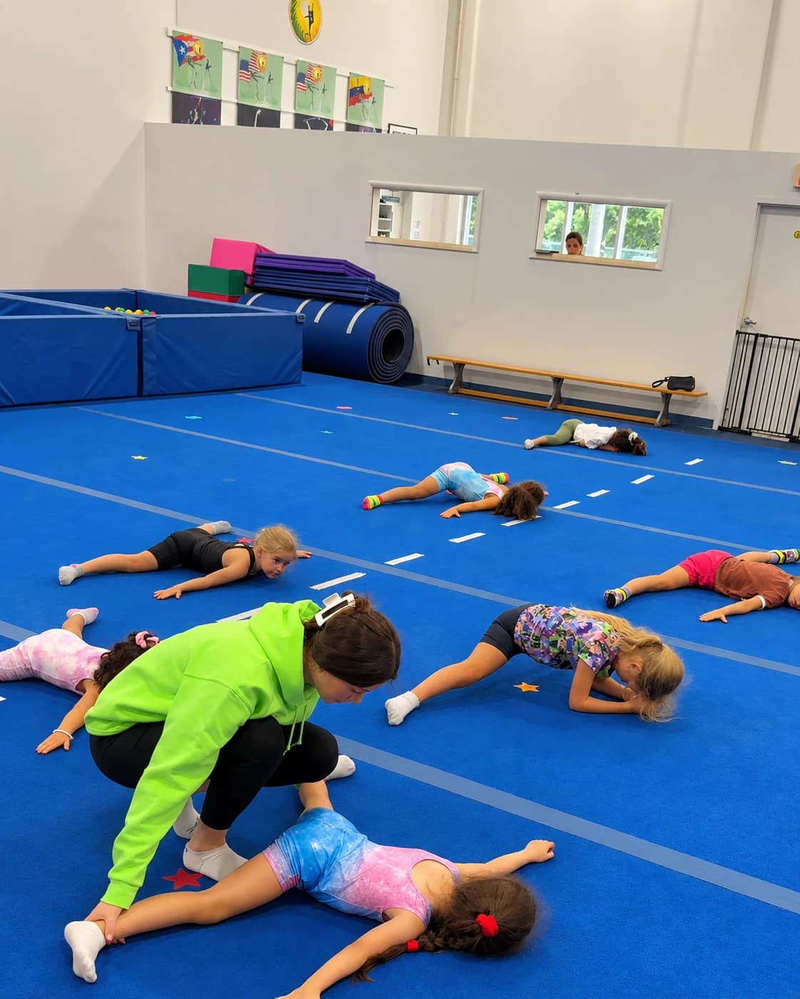 A coach kneels to guide a girl's stretch while the class stretches on the blue floor at Rhythmic Art