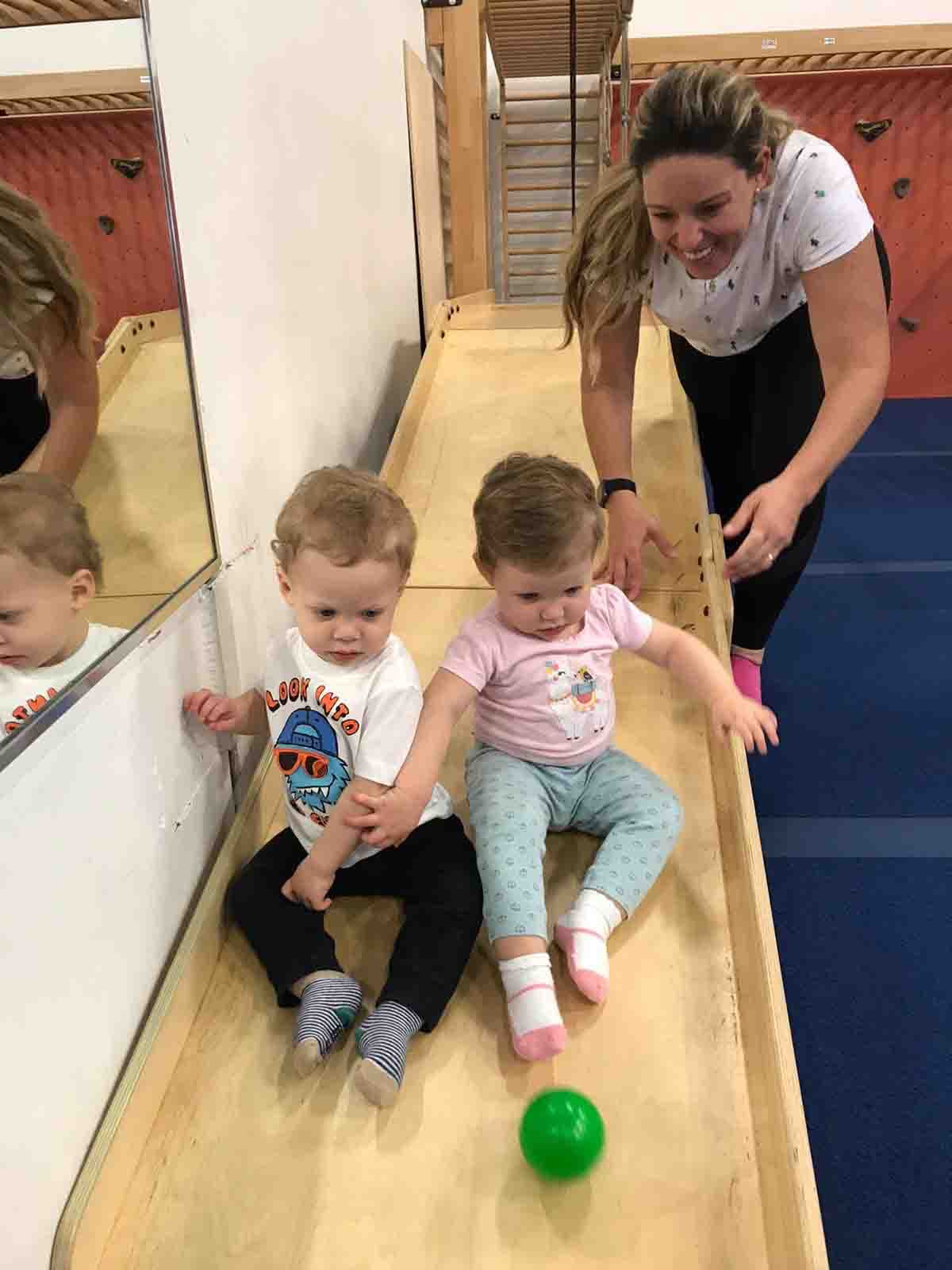 A parent laughs with two toddlers as they play on a wooden ramp in the blue gym