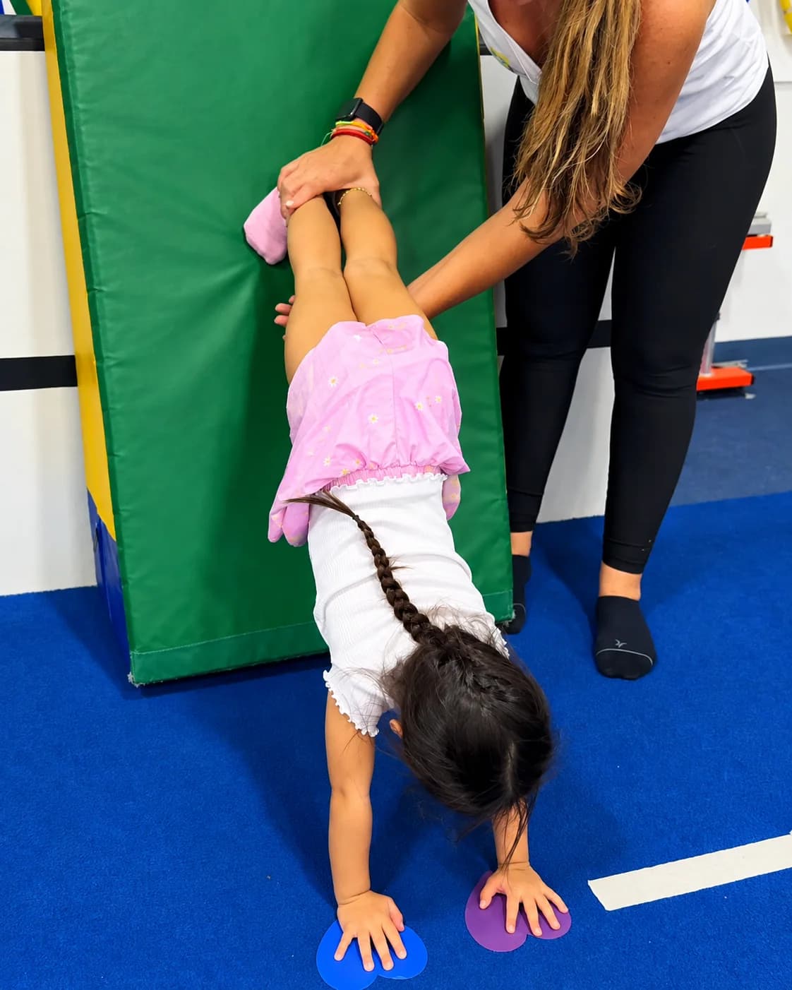 A coach holding a toddler's legs during a handstand on a soft mat at Rhythmic Art